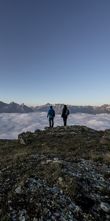 Über den Wolken – Sonnenaufgangswanderung auf den Mot da l’Hom im Unterengadin (Foto: © Andrea Badrutt) Zwei Wanderer stehen auf dem Mot da l’Hom über einem Wolkenmeer mit Blick auf die Unterengadiner Bergwelt