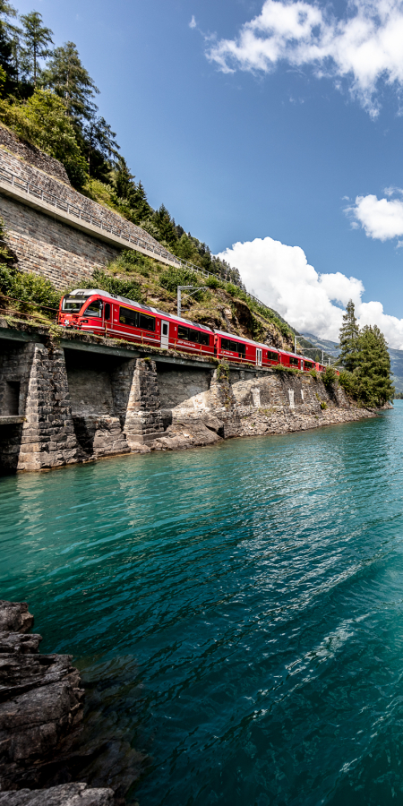 Zwischen Wasser und Bergen: Die Rhätische Bahn folgt dem Ufer des Lago di Poschiavo Richtung Tirano (Foto: © Rhätische Bahn / Andrea Badrutt) Regionalzug der Rhätischen Bahn am Lago di Poschiavo an einem sonnigen Sommertag