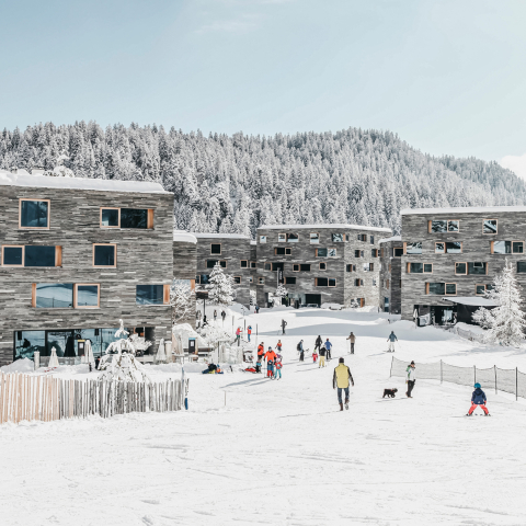 Direkt an der Talstation gelegen: Im rocksresort in Laax beginnt das Skivergnügen gleich vor der Haustür – modernes Design trifft auf alpines Lebensgefühl (Foto: © LAAX / Weisse Arena Gruppe) Winterliche Ansicht des rocksresort in Laax mit Skifahrenden vor moderner Architektur und verschneitem Wald