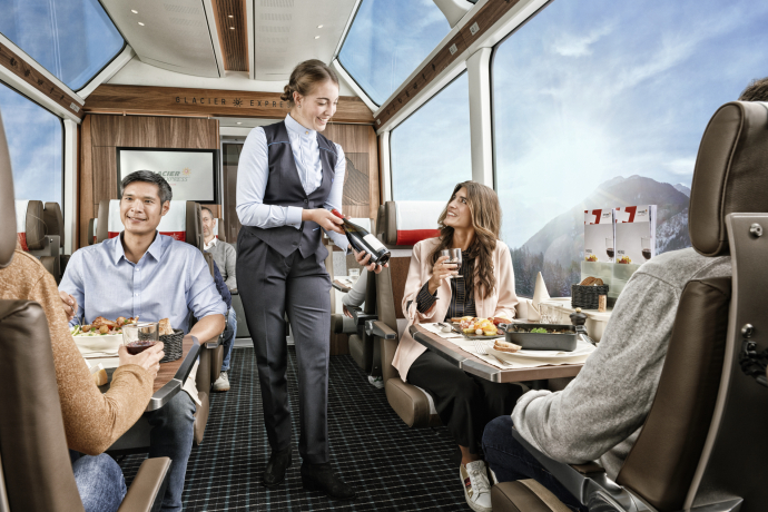 Glacier Express (Foto: © Glacier Express AG / Stefan Schlumpf) Eine Zugbegleiterin zeigt einer Frau im Glacier Express eine Weinflasche