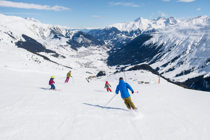 Skifahren mit Panoramablick: Abfahrt in der SkiArena Andermatt-Sedrun (Foto: © Valentin Luthiger) Skifahrer*innen fahren bei Sonnenschein eine breite Piste in der SkiArena Andermatt-Sedrun hinunter, im Hintergrund ein verschneites Tal und umliegende Berglandschaft
