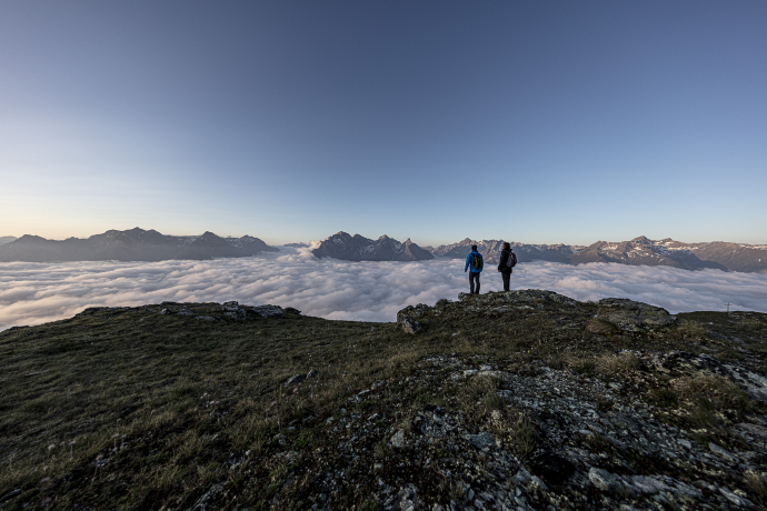 Über den Wolken – Sonnenaufgangswanderung auf den Mot da l’Hom im Unterengadin (Foto: © Andrea Badrutt) Zwei Wanderer stehen auf dem Mot da l’Hom über einem Wolkenmeer mit Blick auf die Unterengadiner Bergwelt